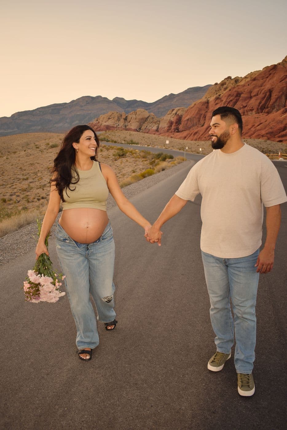 An expecting couple holding hands and smiling at each other on a quiet road below the red cliffs of Red Rock Canyon at sunset
