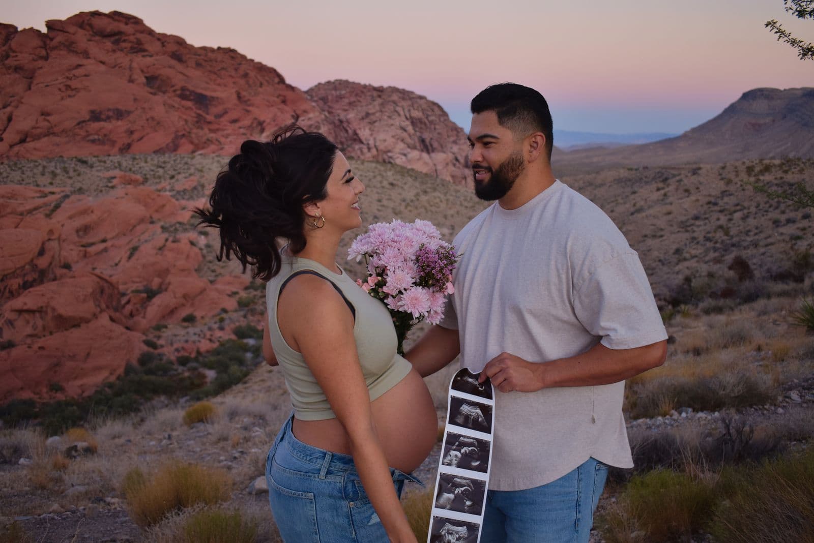An expecting couple holding an ultrasound strip and a bunch of flowers below the red cliffs of Red Rock Canyon at dusk